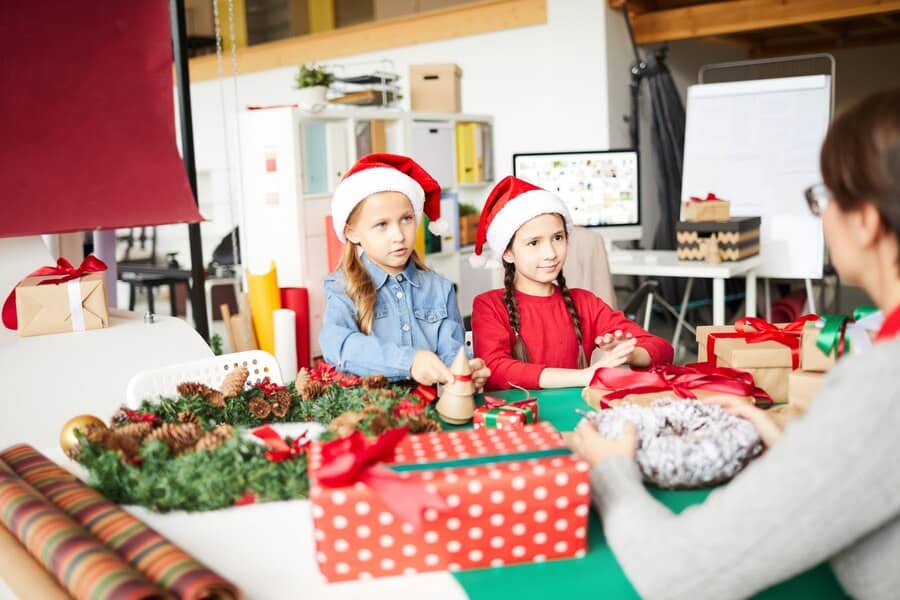 Two children joyfully sit at a table surrounded by colorful Christmas presents at school, celebrating the festive season.