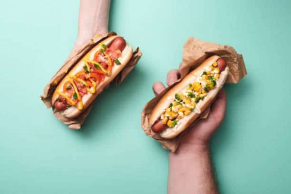 cropped view of man and woman holding two tasty hot dogs in paper on blue